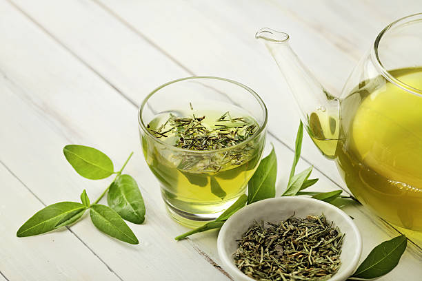 A glass of fresh green tea with visible tea leaves, accompanied by a teapot and loose green tea leaves on a light wooden surface.